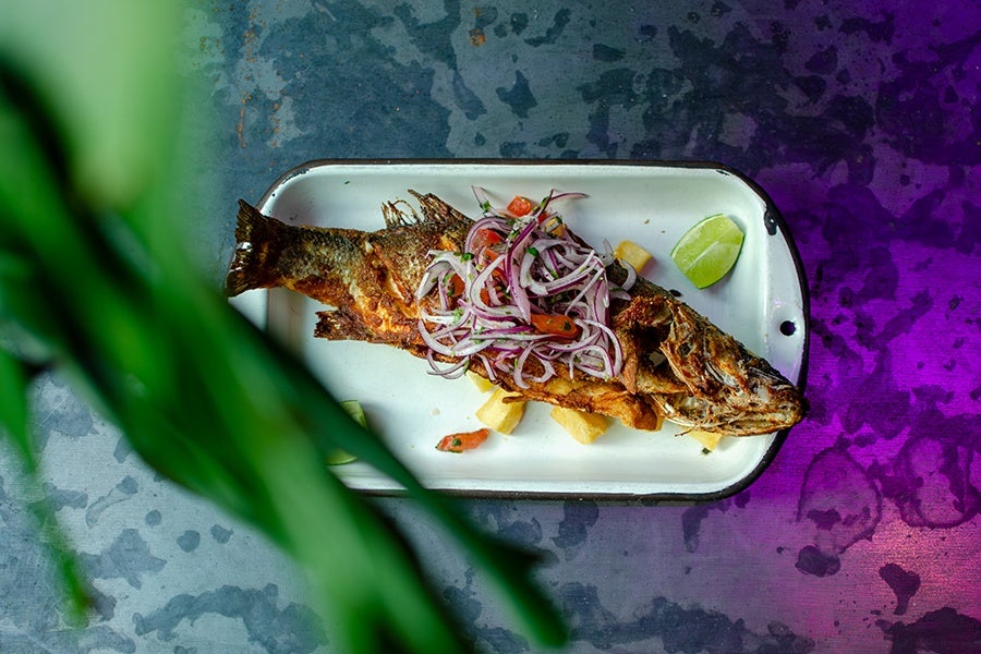 A whole fried fish served on a white rectangular plate, topped with a fresh salad of sliced red onions and tomatoes, accompanied by fried potato pieces and a lime wedge on the side. The plate is set on a textured surface with a green plant partially visible in the foreground.