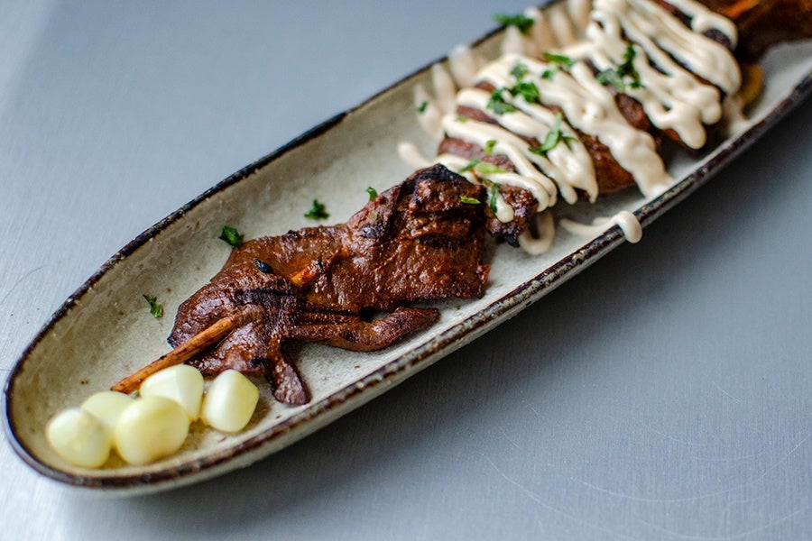 Grilled meat skewer drizzled with a creamy white sauce and garnished with chopped herbs, served on a long, oval ceramic plate with a few pieces of corn on the cob at one end.