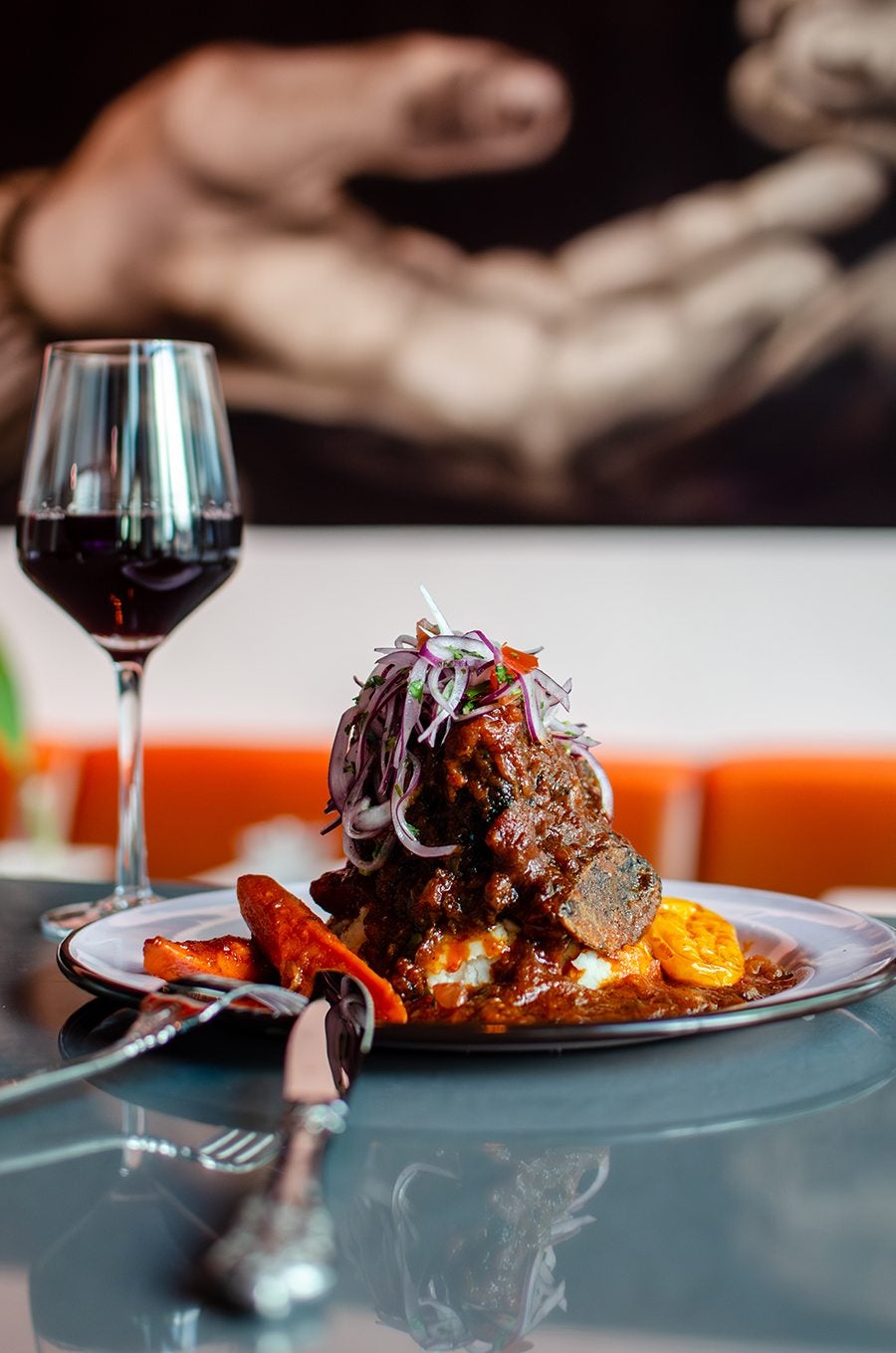 A plated dish featuring a rich, dark stew with chunks of meat, topped with thinly sliced red onions and herbs, served alongside roasted carrots and a yellow vegetable. A glass of red wine is placed to the left of the plate, with a fork and knife resting on the table in front. The background shows a blurred image of hands and an orange seating area.
