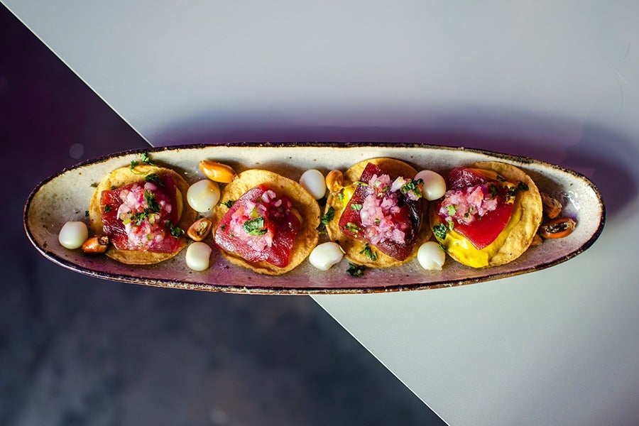 Four small round appetizers on a long oval ceramic plate, each topped with diced red fish, chopped onions, and herbs, served on crispy golden bases with scattered corn kernels and white beans around them.