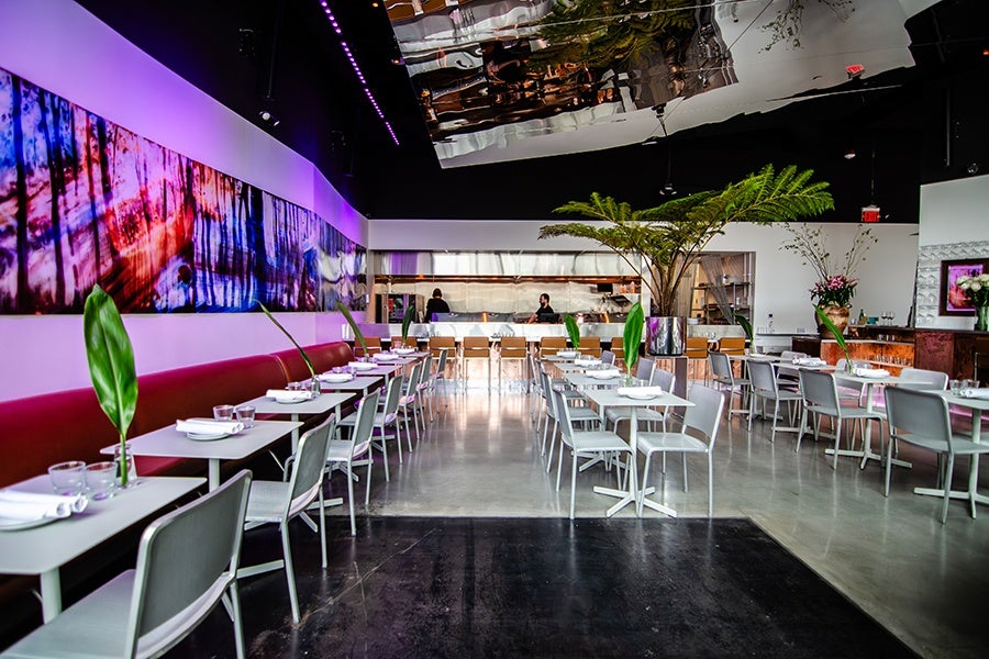 Modern restaurant interior with white tables and gray chairs arranged in rows. A long red cushioned bench lines the left wall, which features a large colorful abstract painting illuminated by purple lighting. The ceiling has a reflective metallic surface, and there are large green plants in vases placed on some tables. The back of the room shows an open kitchen area with two staff members visible. The floor is polished concrete.