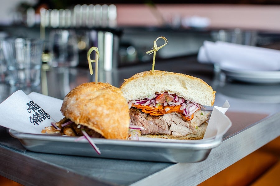 A sandwich cut in half on a metal tray lined with paper. The sandwich contains sliced meat, shredded red onions, and thin slices of orange vegetables, all inside a soft, crusty white bread roll. Each half is secured with a bamboo skewer. The tray is placed on a gray table with blurred background elements including glasses and plates.