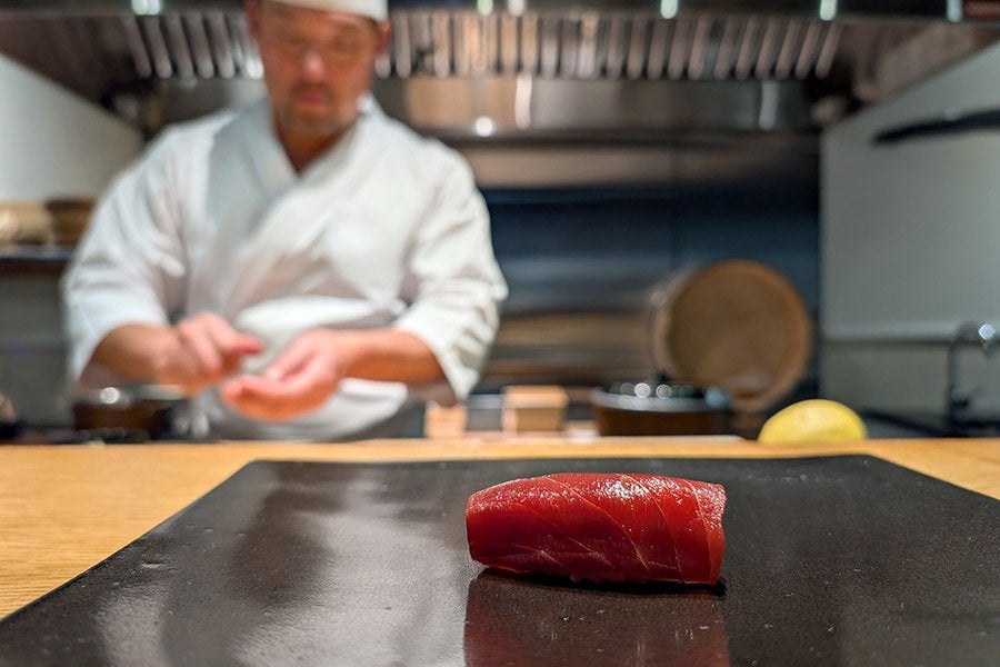 A piece of red tuna sushi placed on a black rectangular plate, with a sushi chef in a white uniform and hat blurred in the background preparing food. The setting appears to be a sushi bar or restaurant.
