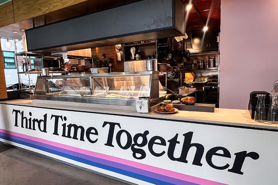 A modern café counter with a glass display case, wooden countertop, and a sign on the front reading "Third Time Together" in large black letters. The counter has a striped design in pink, purple, and blue near the bottom. Behind the counter, there are kitchen shelves, utensils, and equipment, with some pastries placed on the counter. The setting has a warm, inviting atmosphere with wooden and metal elements.