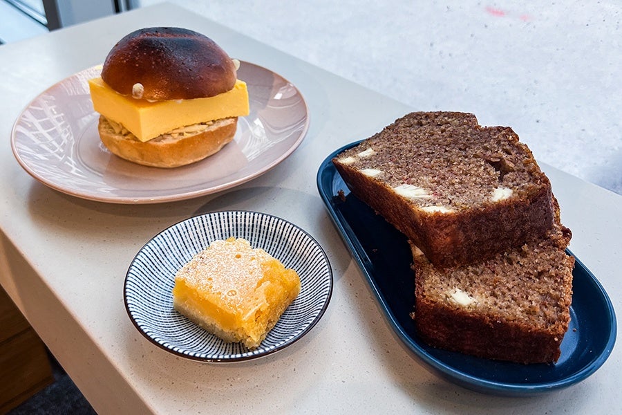 A plate with a sandwich featuring a thick slice of yellow cheese and a bun, a small dish with a square lemon bar dusted with powdered sugar, and a blue plate holding two slices of dark brown bread with chunks of cheese inside.