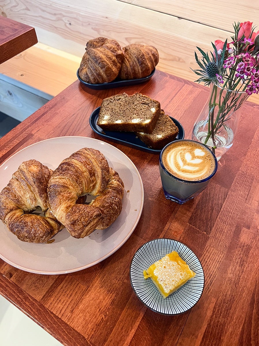 Two croissants on a beige plate, two more croissants on a black tray, two slices of brown bread with white chunks on another black tray, a glass of latte with leaf latte art, a small square yellow pastry dusted with powdered sugar on a small round plate, and a glass vase with pink and purple flowers on a wooden table.
