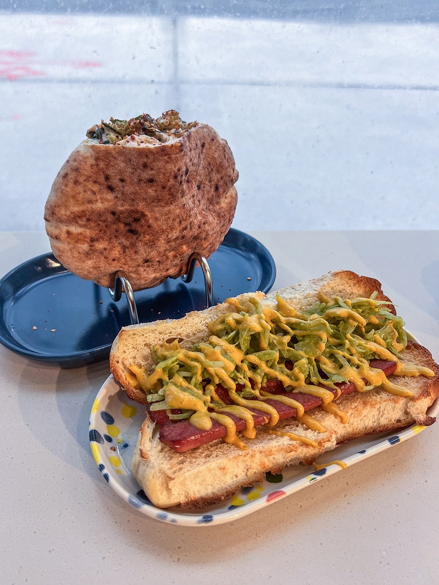 A pita sandwich filled with vegetables and sauce is held upright on a metal stand on a dark blue plate. In front, an open sandwich on toasted bread features a sausage topped with shredded lettuce and drizzled with mustard, served on a colorful, patterned plate.
