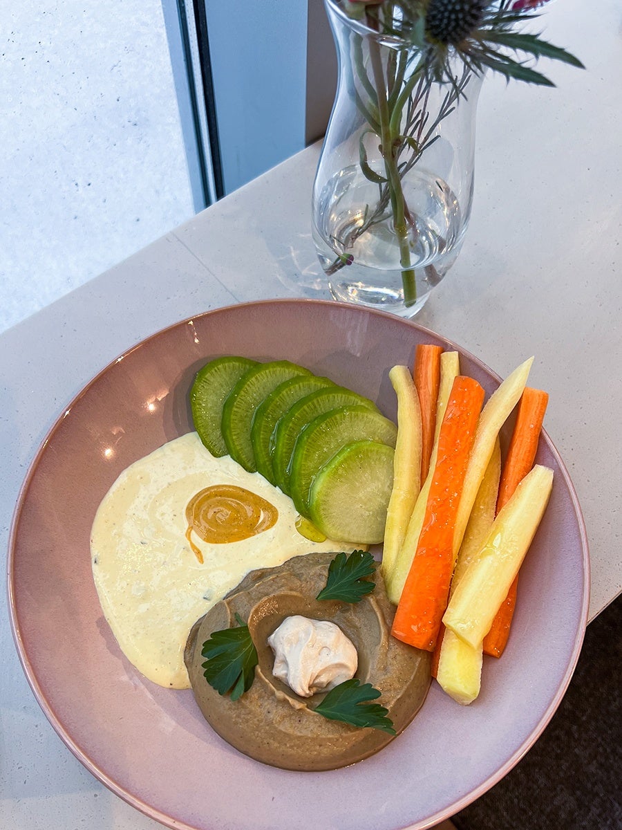 A pink plate with a serving of creamy dips, one light beige with a swirl of mustard and the other brown topped with a dollop of white sauce and garnished with parsley leaves. Next to the dips are sliced green cucumbers and sticks of orange and yellow carrots. A clear glass vase with water and flowers is in the background on a white countertop.
