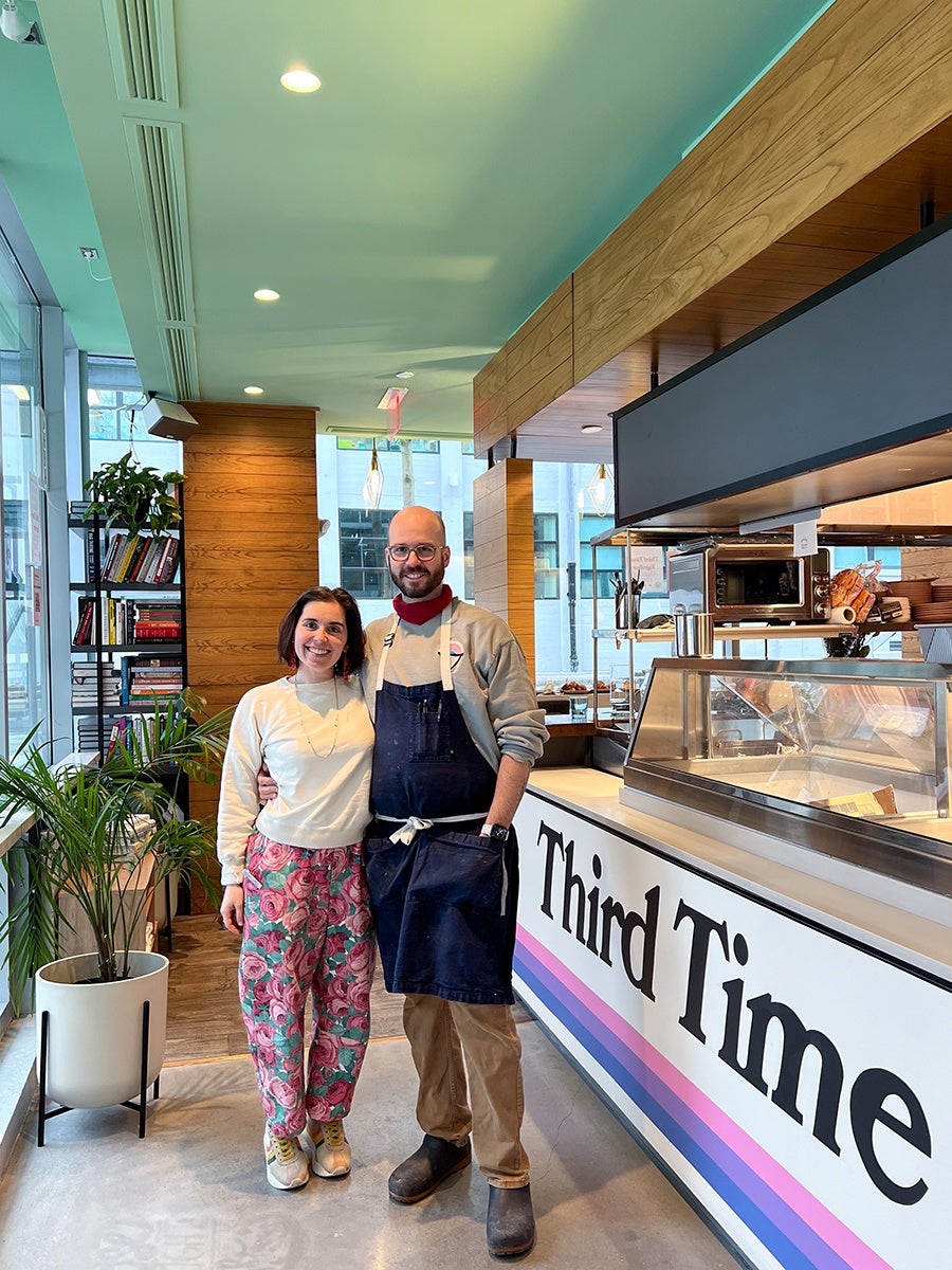A smiling man and woman stand side by side inside a modern cafe or bakery. The man wears glasses, a beige sweatshirt, a dark blue apron, tan pants, and dark shoes. The woman wears a white sweatshirt, colorful floral pants, and white sneakers. Behind them is a counter with a glass display case and a sign that reads "Third Time" with pink, purple, and blue stripes below the text. The interior features wood paneling, large windows, potted plants, and a bookshelf filled with books.