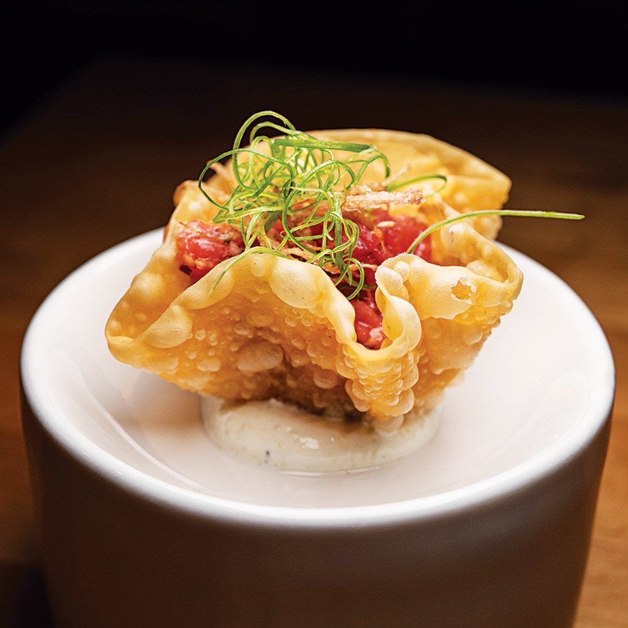 A crispy, golden-brown wonton cup filled with diced raw tuna and topped with thin, curly green garnish, served on a small dollop of creamy white sauce in a white ceramic dish.