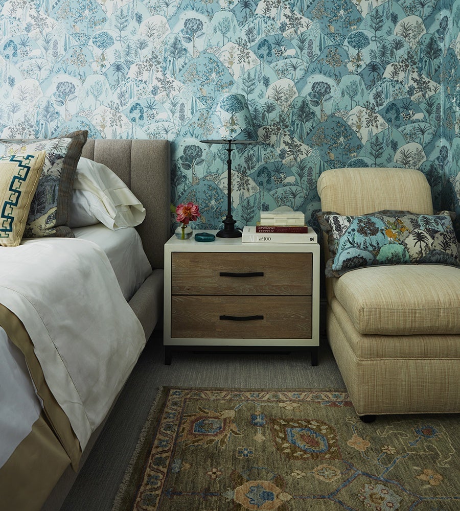 A cozy bedroom corner featuring a beige upholstered bed with white and beige bedding, a two-drawer nightstand with a white frame and wooden drawer fronts, and a beige cushioned chair. The nightstand holds a black lamp with a blue floral lampshade, a small vase with a pink flower, a blue decorative item, and a stack of books. The wall behind is covered in blue and white wallpaper with a botanical pattern, and a patterned rug with earthy tones lies on the floor. The chair has a decorative pillow with a matching blue botanical design.