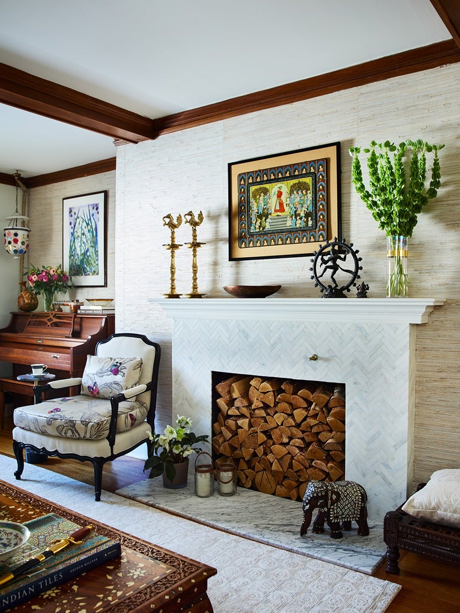 A cozy living room corner featuring a white marble fireplace filled with stacked firewood. On the mantel are decorative items including two brass candle holders, a wooden bowl, a black statue of a dancing figure inside a circular frame, and a tall glass vase with green leafy stems. Above the mantel hangs a framed colorful painting with a traditional theme. To the left of the fireplace is an upholstered armchair with floral cushions, a small side table with a cup and saucer, and a wooden piano with a vase of pink flowers on top. The floor has a light-colored rug, and a carved wooden elephant figurine sits on the hearth. The room has wooden beams on the ceiling and a framed floral artwork on the wall near the piano.
