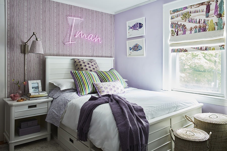 Bedroom with a white wooden bed frame and headboard, adorned with multiple pillows in purple, green, and striped patterns. A purple throw blanket is draped across the bed. The wall behind the bed features vertical striped wallpaper and a pink neon sign spelling "Iman." Next to the bed is a white nightstand with books, a framed photo, a lamp, and decorative items. The adjacent wall is painted light purple and has two framed fish illustrations. A window with a colorful patterned Roman shade lets in natural light. Two woven baskets with lids are placed near the window.