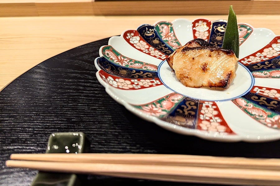 A small piece of fish garnished with a green vegetable, served on a decorative plate with red, blue, green, and white patterns. The plate is placed on a black textured tray, with wooden chopsticks resting on a green chopstick holder in the foreground.