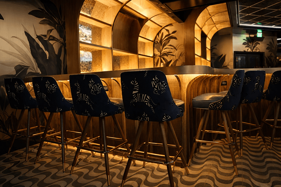A stylish bar area with a curved wooden counter illuminated by warm, recessed lighting. The bar stools have dark blue upholstery featuring a zebra pattern and wooden legs with gold footrests. The floor has a wavy, patterned design, and the walls are decorated with large botanical murals in muted tones.