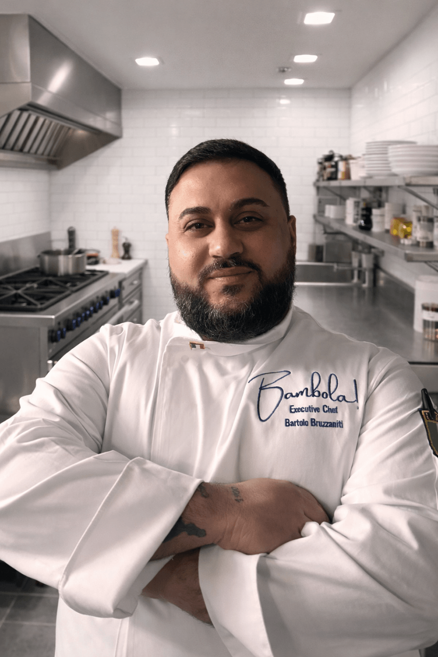 A bearded man wearing a white chef's coat with the embroidered text "Bambola Executive Chef Bartolo Bruzzaniti" stands confidently with arms crossed in a professional kitchen. The kitchen features stainless steel appliances, white tiled walls, and shelves with plates and various containers.